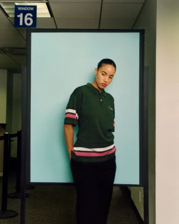 An adult female stands in a casual pose in an office setting with a blank blue sign and dark green polo shirt with white and pink stripes.