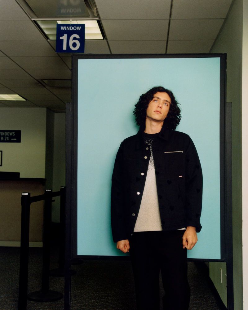 Full-body portrait of a young adult man with curly hair in a black jacket and pants standing in front of a light blue wall indoors.
