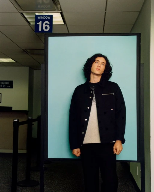 Full-body portrait of a young adult man with curly hair in a black jacket and pants standing in front of a light blue wall indoors.