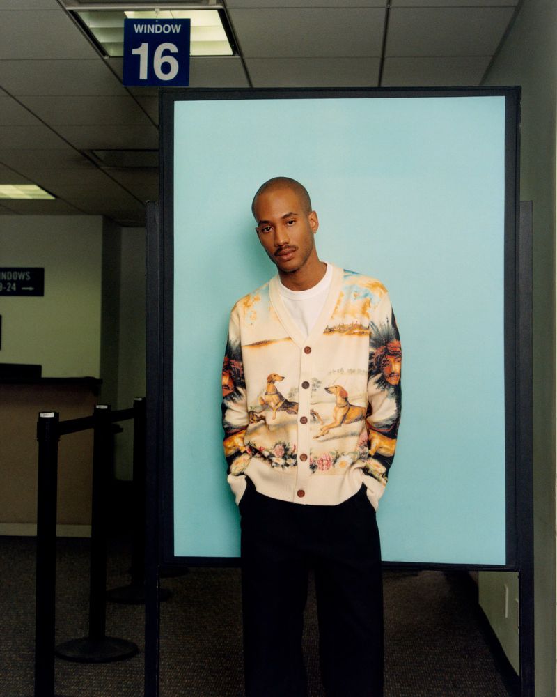 Adult male wearing a patterned cardigan in a brightly lit indoor space with blue accent wall and dark trim in a medium shot.