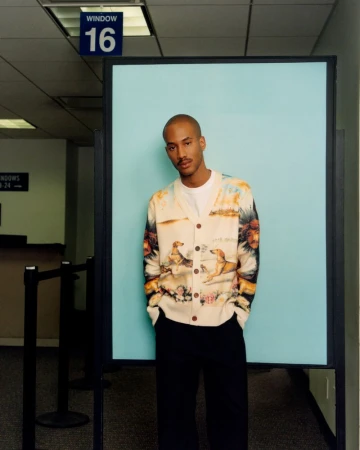 Adult male wearing a patterned cardigan in a brightly lit indoor space with blue accent wall and dark trim in a medium shot.