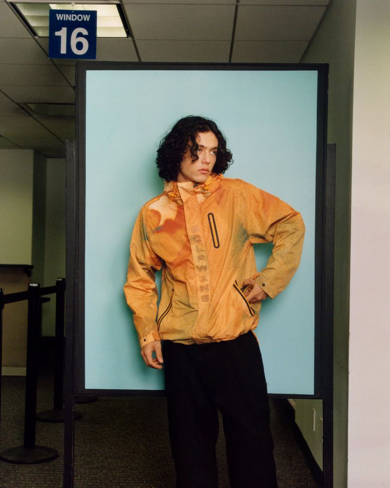 Young adult in an orange jacket and black pants standing in front of a blue wall with a 'Window 16' sign above it.