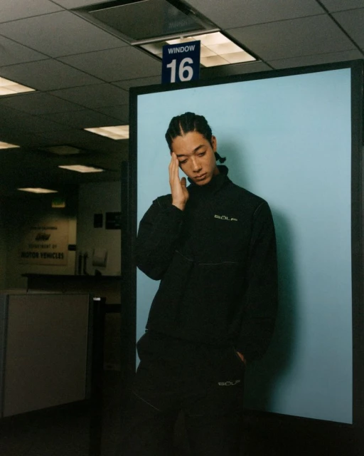 Asian man with hand to head in black athletic wear stands in front of light blue wall in indoor waiting room with overhead lighting.