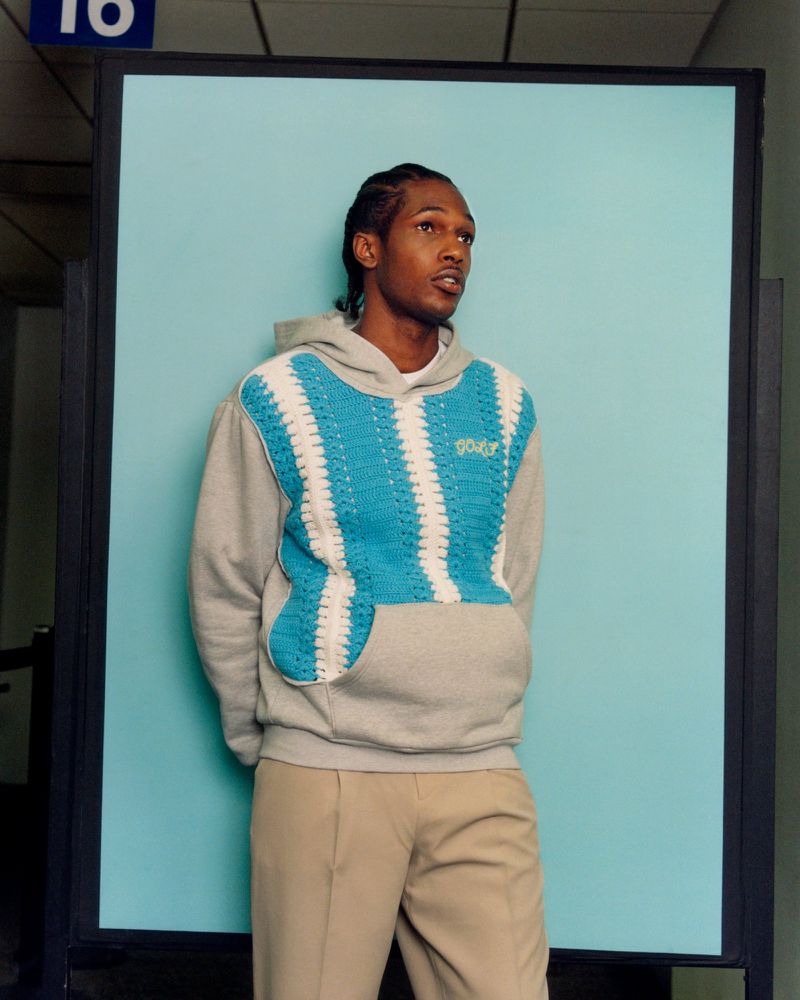 Adult male poses in a grey and turquoise hoodie and khaki pants in an indoor environment with a light blue background.