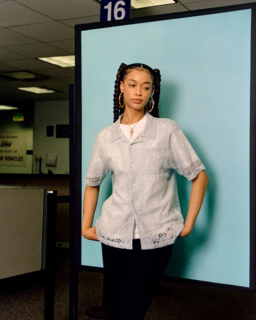 Portrait of a young woman with braided pigtails wearing a sheer blue lace top in what appears to be a DMV or government office.