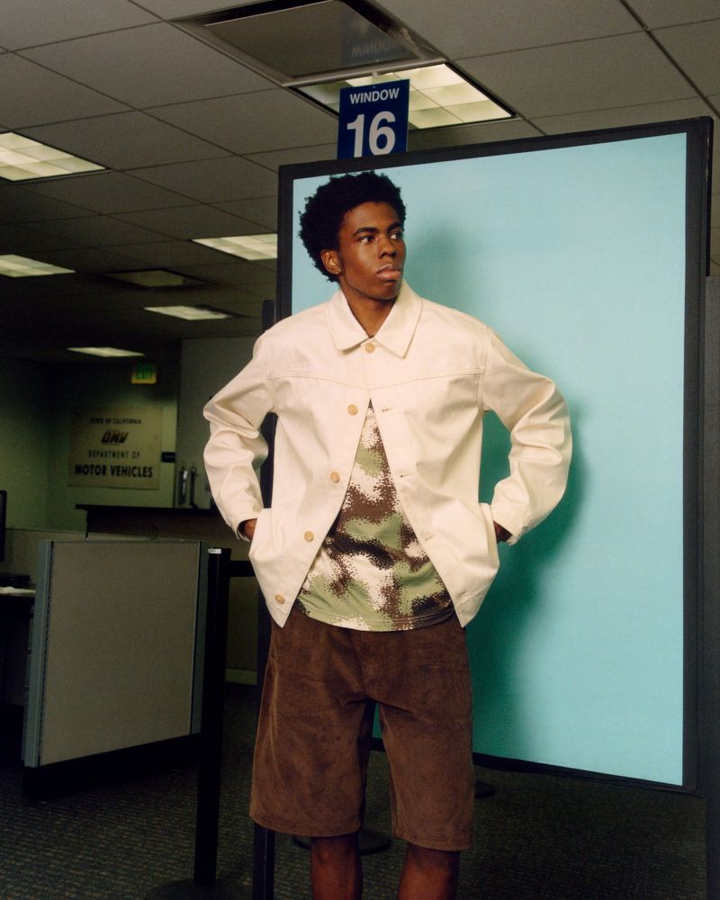 Young adult man in a cream jacket and brown shorts stands in front of window 16 at the Department of Motor Vehicles.