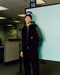 Model wearing a black beanie, green hoodie, and black vest in a DMV office setting for Golf Wang S/S 2024.