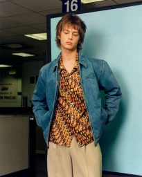 Model in a blue denim jacket, patterned photo-print shirt, and beige trousers standing in a DMV office setting.