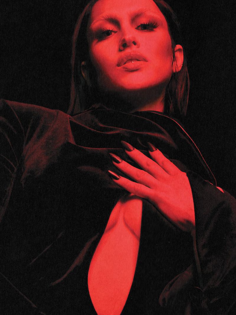 Portrait of a young adult female model with dark nails, pale eyebrows and lips, wearing a low-cut black top, against a dark background.
