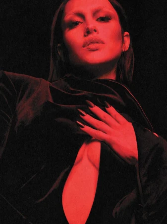 Portrait of a young adult female model with dark nails, pale eyebrows and lips, wearing a low-cut black top, against a dark background.