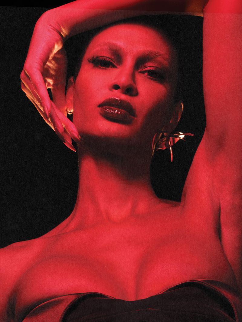 Portrait of a woman with red skin and dark lipstick, wearing a black strapless top against a dark background, illuminated by red lighting.