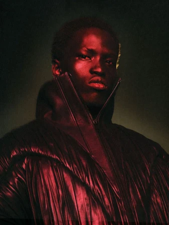 Striking portrait of a Black male fashion model in a reflective maroon puffer jacket, dramatic lighting, and a blurred olive green background.