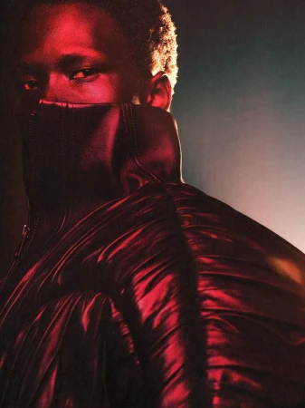 Close-up of a Black male model in a red padded jacket with the collar pulled up over the lower half of his face, dark background.