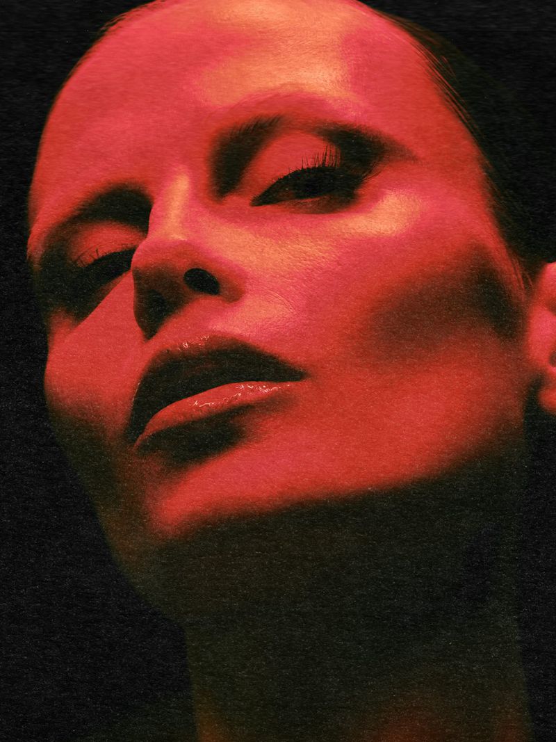 Close-up portrait of an adult female with fair skin in a low angle shot, bathed in vibrant red lighting against a dark, grainy background.