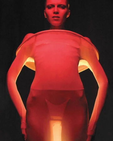 Full body shot of a slim adult female wearing a translucent pink bodysuit with orange glow against a dark background in a studio.