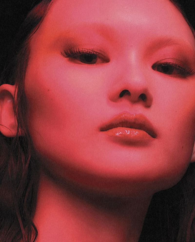 Close-up beauty shot of an adult female with fair skin, wearing glossy lip color, and dramatic eye makeup, with a dark background.