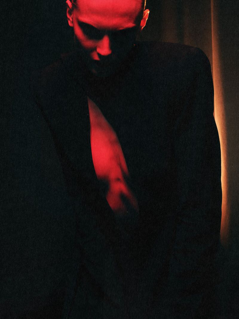Model wearing an open structured black blazer illuminated by dramatic red light against a dark background.