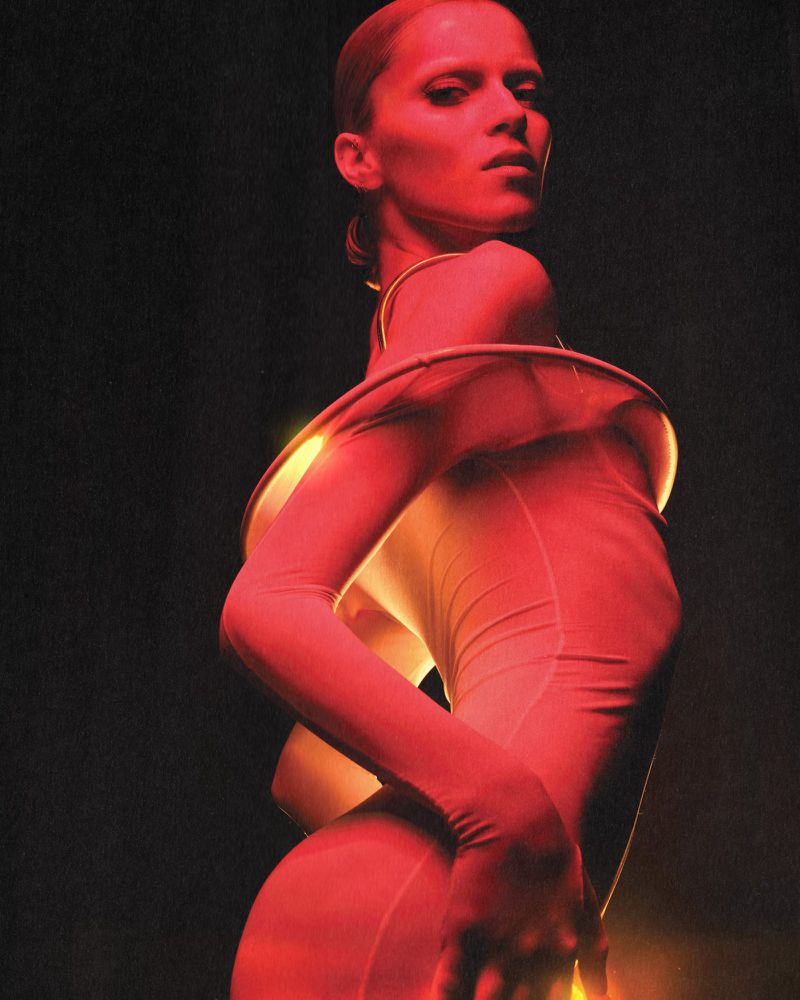 A poised woman in a fitted outfit with draped accents stands against a dark background, bathed in red and golden light, highlighting her form.