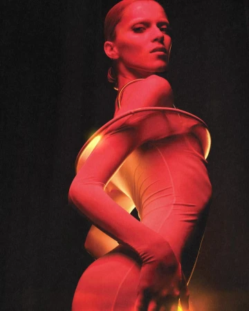 A poised woman in a fitted outfit with draped accents stands against a dark background, bathed in red and golden light, highlighting her form.