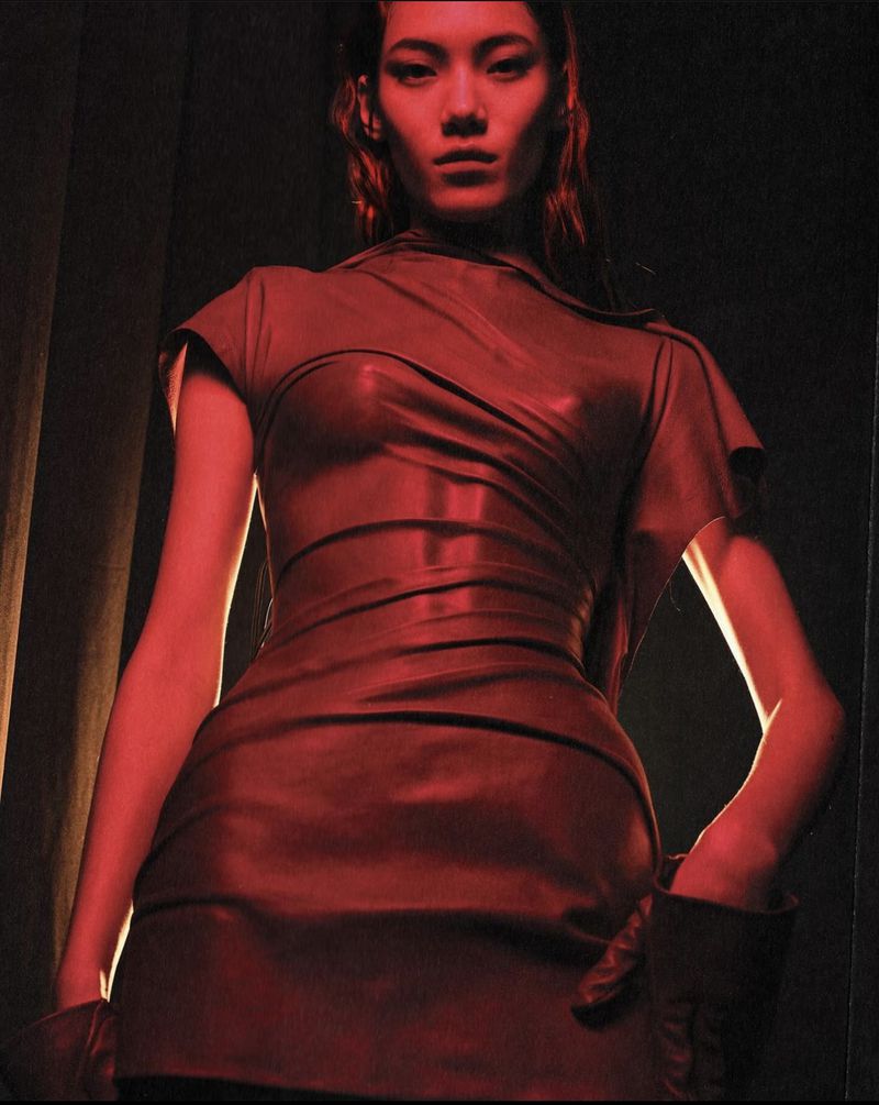 Asian woman in red latex dress and gloves gazes down from an angle with dramatic lighting and dark background in a fashion editorial style.