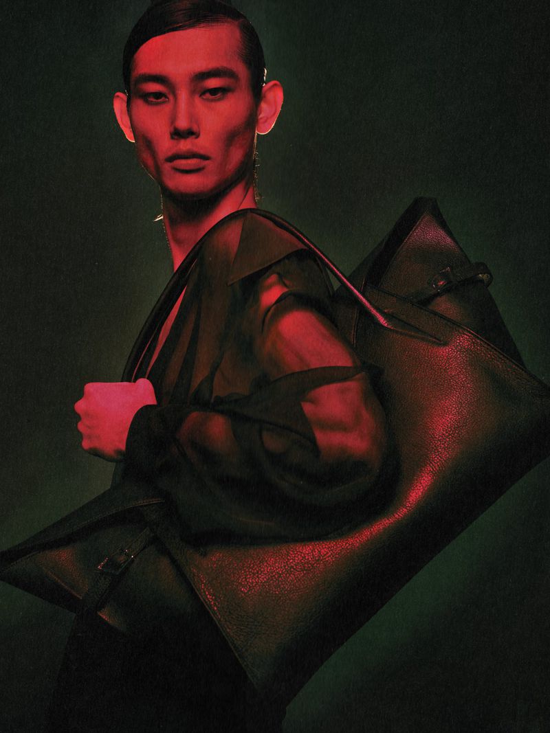 Asian male fashion model poses with a large black leather bag in a studio with red and green lighting.
