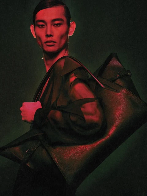 Asian male fashion model poses with a large black leather bag in a studio with red and green lighting.
