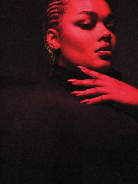 Close-up portrait of an adult female with cornrow braids, hand on neck, wearing a black turtleneck against a dark backdrop in studio lighting.