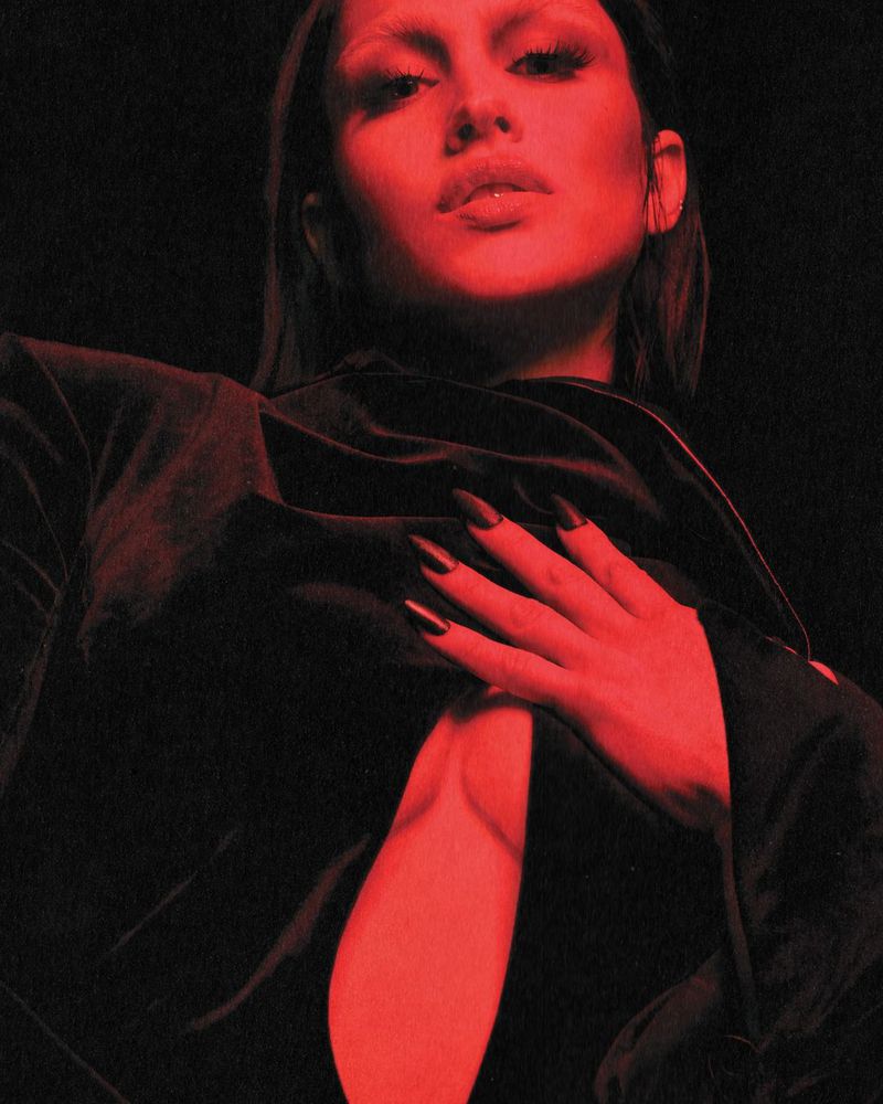 Portrait of a woman with long dark hair, long black nails, wearing a dark top, posing against a dark red background.
