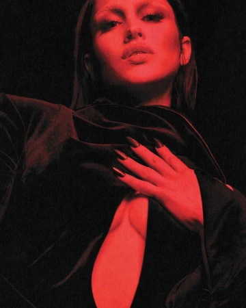 Portrait of a woman with long dark hair, long black nails, wearing a dark top, posing against a dark red background.