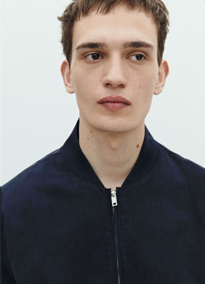 A light-skinned adult male model wearing a blue bomber jacket, captured in a medium shot against a plain white background in a fashion setting.
