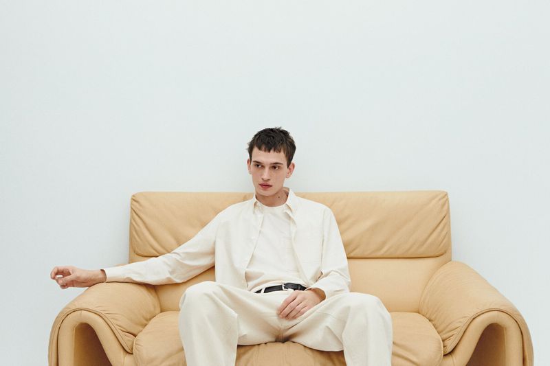Young adult male model in casual off-white shirt and pants sitting on a vintage tan leather couch against a plain white backdrop.