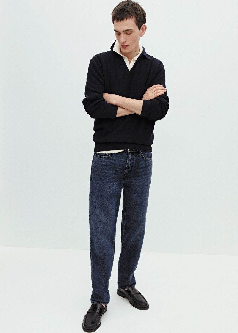 Male model in a black polo sweater and dark wash denim jeans standing with crossed arms against a white studio background.