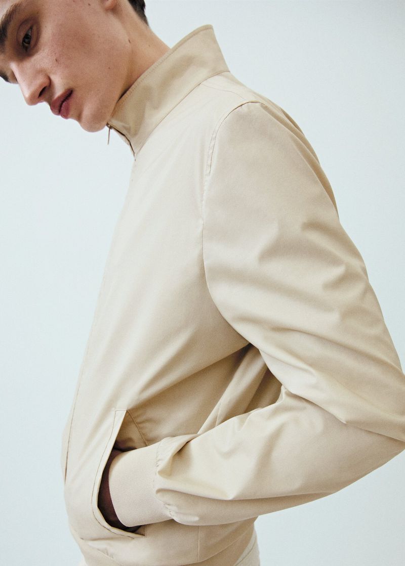 Close-up of a light beige jacket with a high collar and a man wearing it, set against a plain, light-colored background.