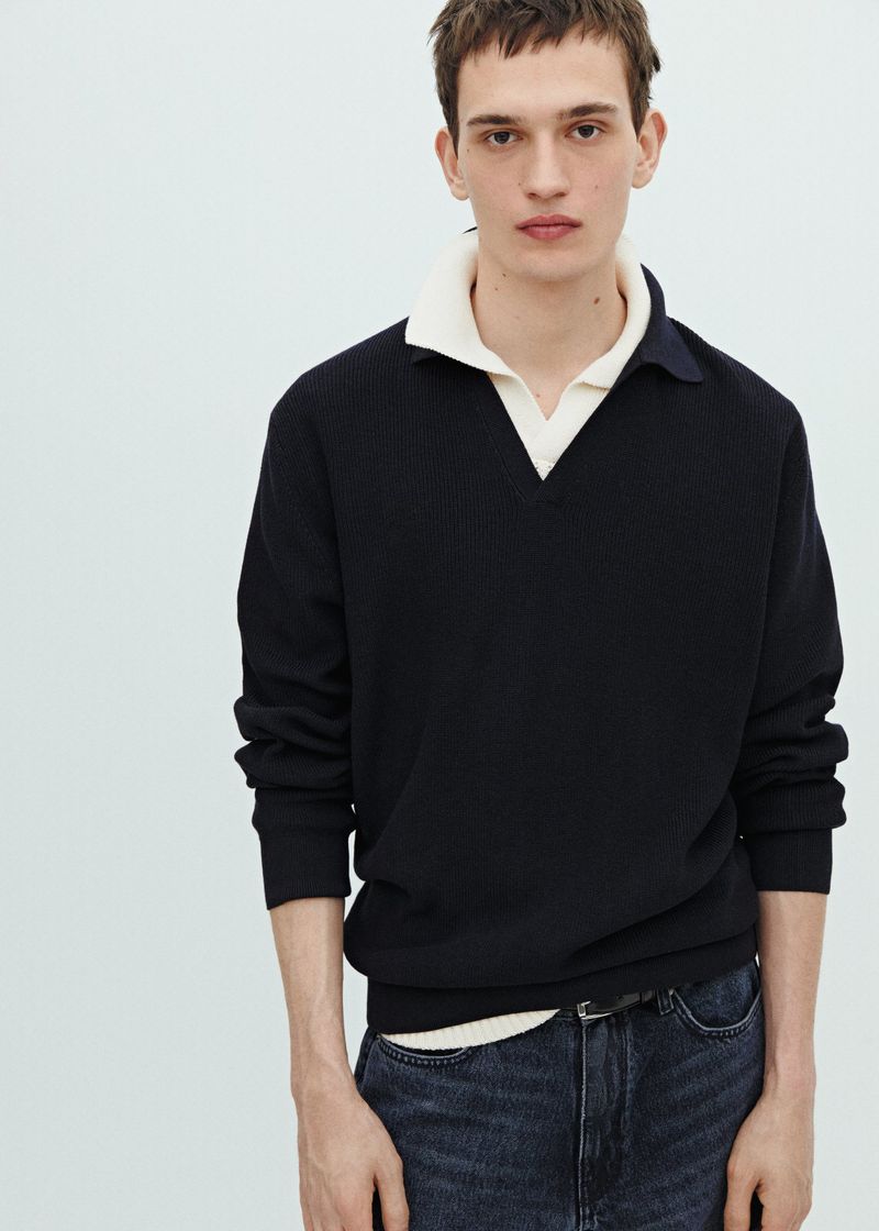 Portrait of a young adult man in a black V-neck sweater with a cream-colored collared shirt underneath and dark jeans against a white background.