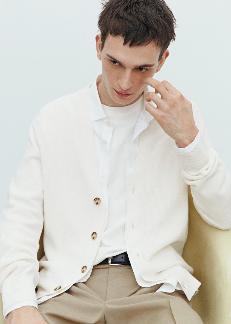A seated male model wearing an off-white cardigan over a white shirt and beige trousers against a plain light background.