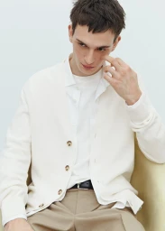 A seated male model wearing an off-white cardigan over a white shirt and beige trousers against a plain light background.
