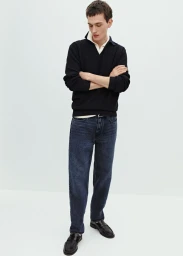 Male model in a black polo sweater and dark wash denim jeans standing with crossed arms against a white studio background.