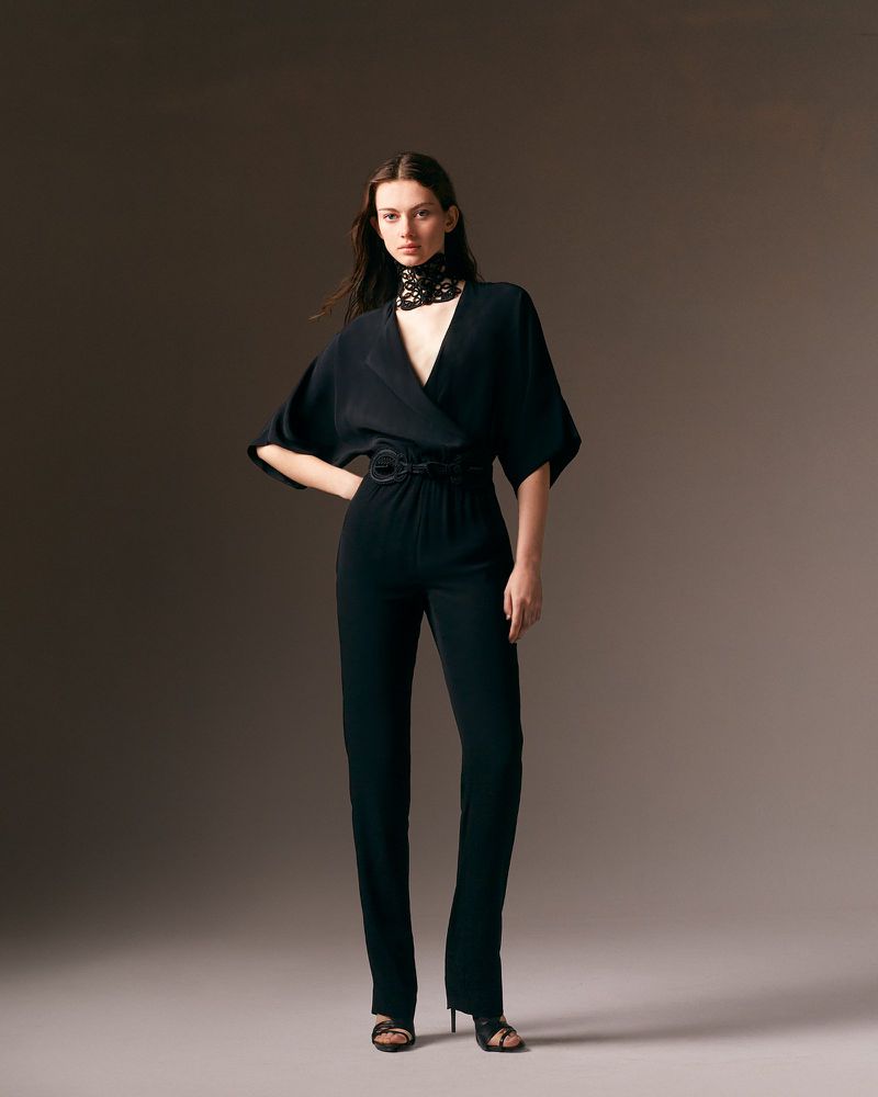 Fashionable woman poses in black jumpsuit with lace collar against a studio backdrop, showcasing contemporary and elegant style.