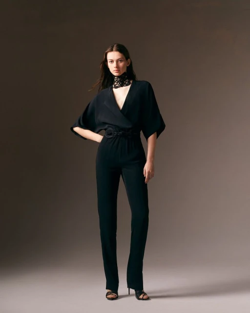Fashionable woman poses in black jumpsuit with lace collar against a studio backdrop, showcasing contemporary and elegant style.