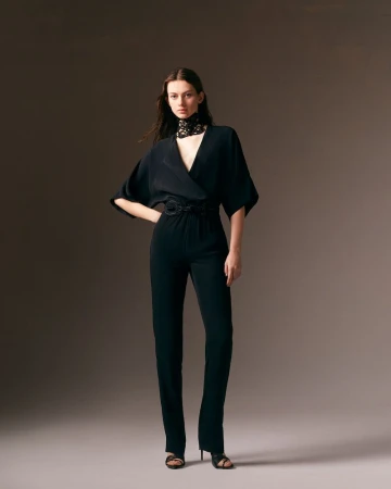 Fashionable woman poses in black jumpsuit with lace collar against a studio backdrop, showcasing contemporary and elegant style.