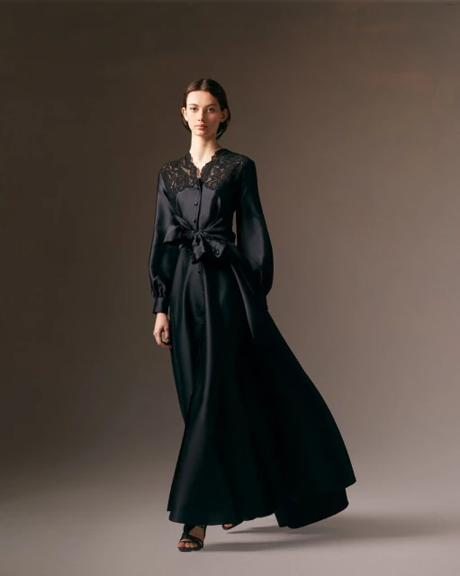 Elegant woman in a flowing black gown with lace details, standing gracefully in a studio with soft, neutral lighting. Fashion portrait.