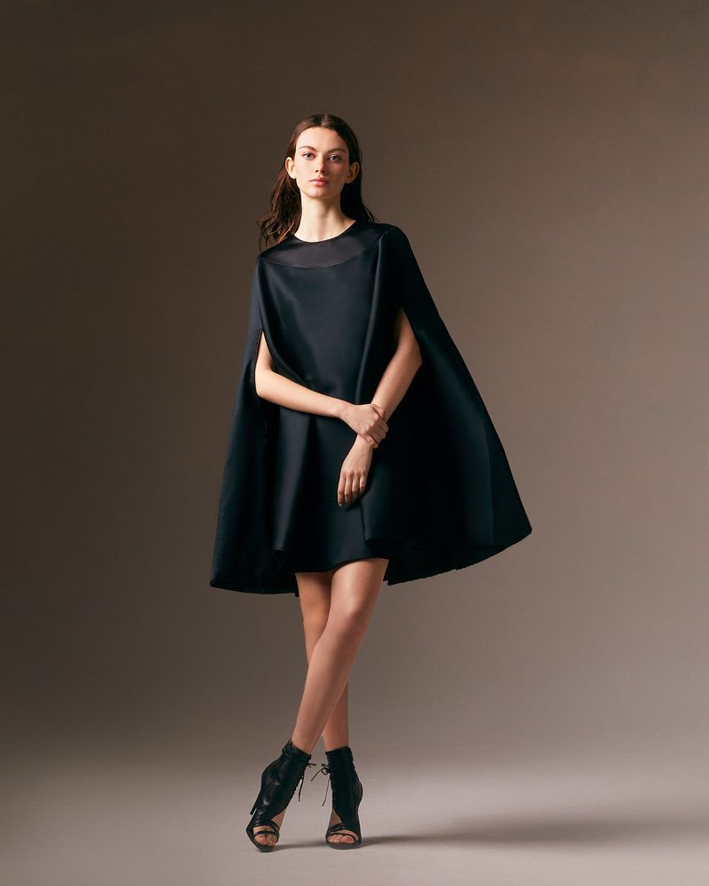 A woman in a short black dress and cape poses in a studio, legs crossed, against a soft, neutral gradient backdrop with subtle lighting.