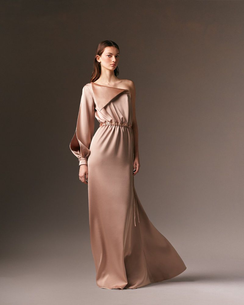 Elegant adult woman posing in a studio wearing a light brown, satin one-shoulder gown with a long sleeve, in a medium studio shot.