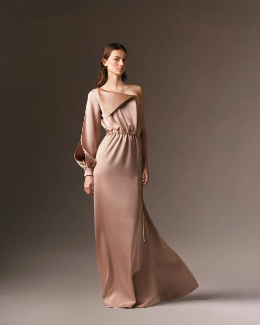 Elegant adult woman posing in a studio wearing a light brown, satin one-shoulder gown with a long sleeve, in a medium studio shot.