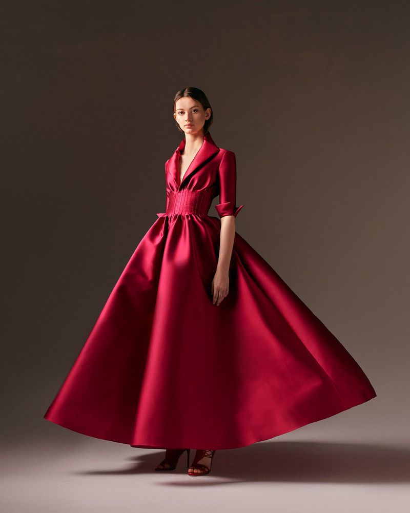 Elegant woman in a voluminous red satin gown with a cinched waist and collar, standing in a neutral setting, fashion portrait.