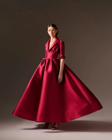 Elegant woman in a voluminous red satin gown with a cinched waist and collar, standing in a neutral setting, fashion portrait.