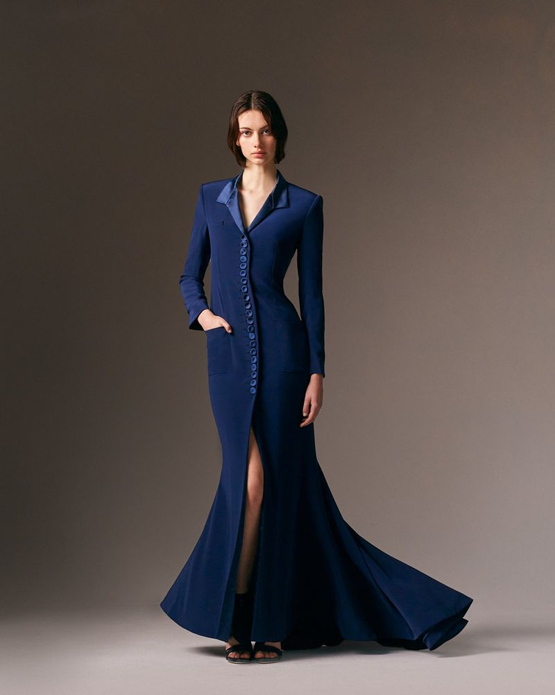 A model poses in a full-length shot wearing a long-sleeved, navy blue gown with button detail and a slit, set against a grey studio backdrop.