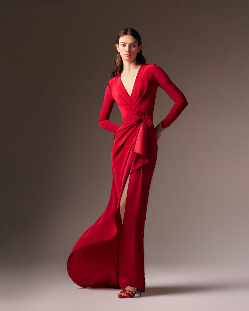 An adult female fashion model poses in a full-length shot wearing a red gown with a side slit, cinched at the waist in a studio setting.