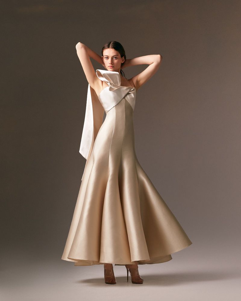 Elegant woman in a champagne-colored gown with a large bow, hands behind her head, stands in a neutral studio.