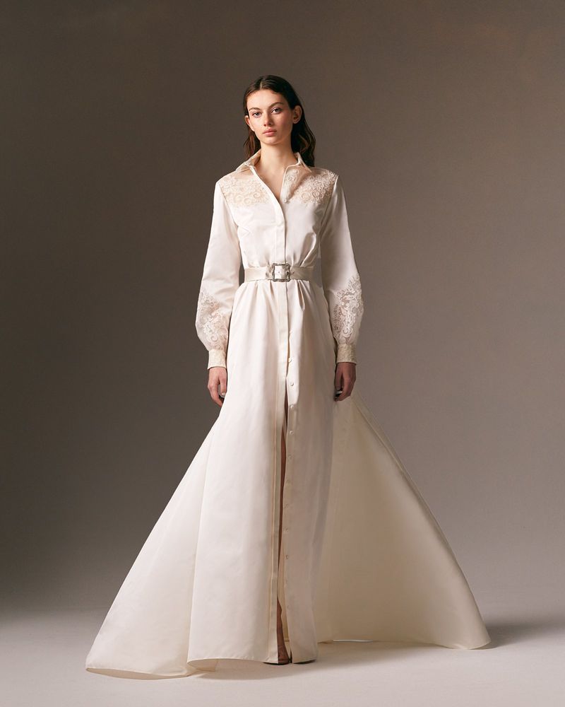 Woman poses in a full-length ivory button-down gown with a long train and lace embellishments against a neutral backdrop in a studio shot.